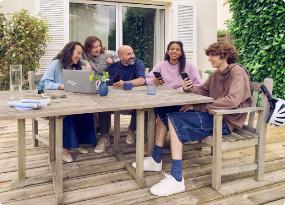 Cinq individus sur leurs ordinateurs portables et smartphones, réunis autour d'une table de jardin sur une terrasse.